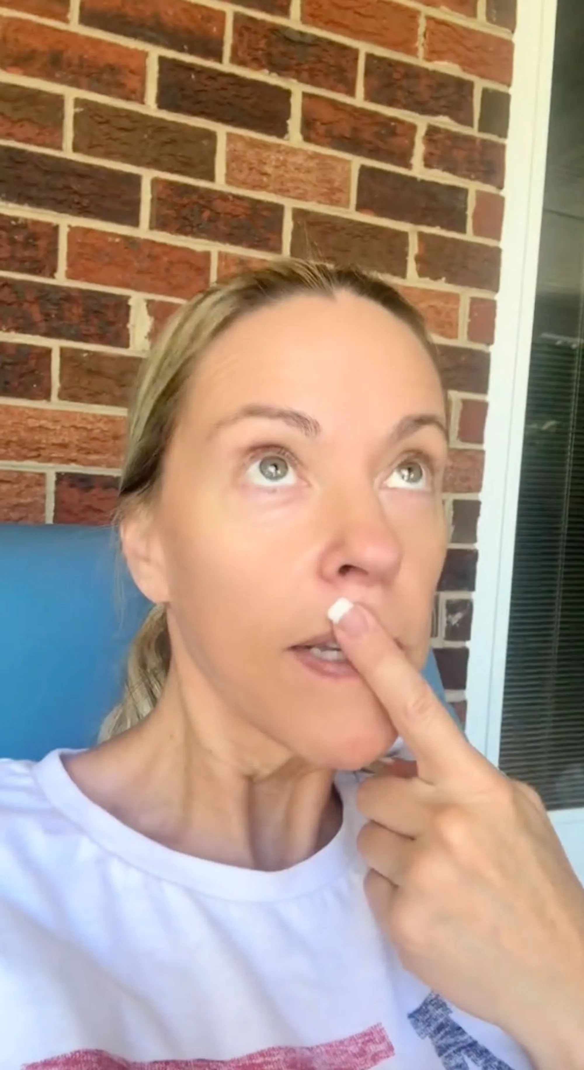 Woman in a white shirt with red and blue stripes looking up and touching her nose with her finger.