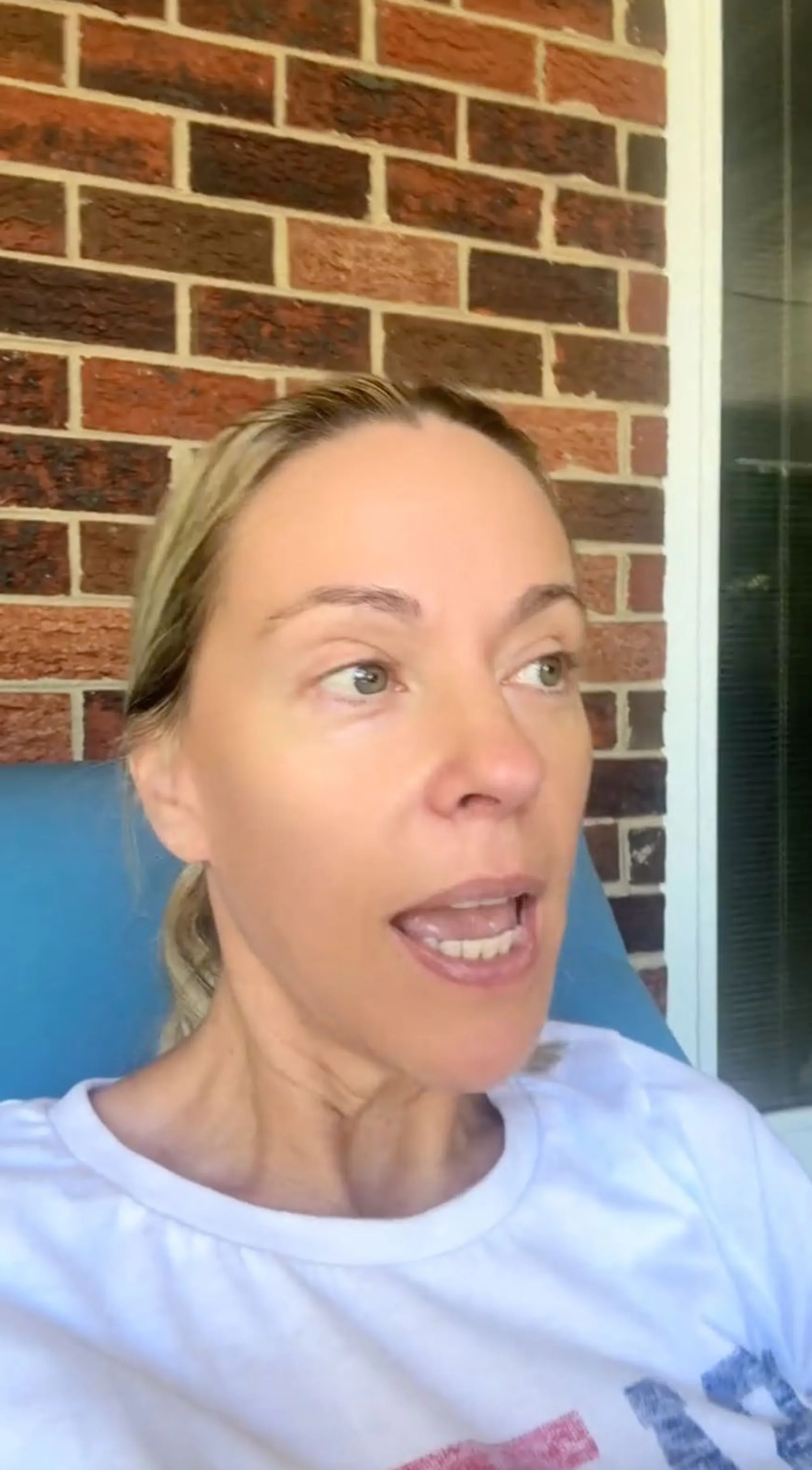 Kate Gosselin with blonde hair pulled back, wearing a white shirt and sitting in a blue chair, speaking to the side with an open mouth.