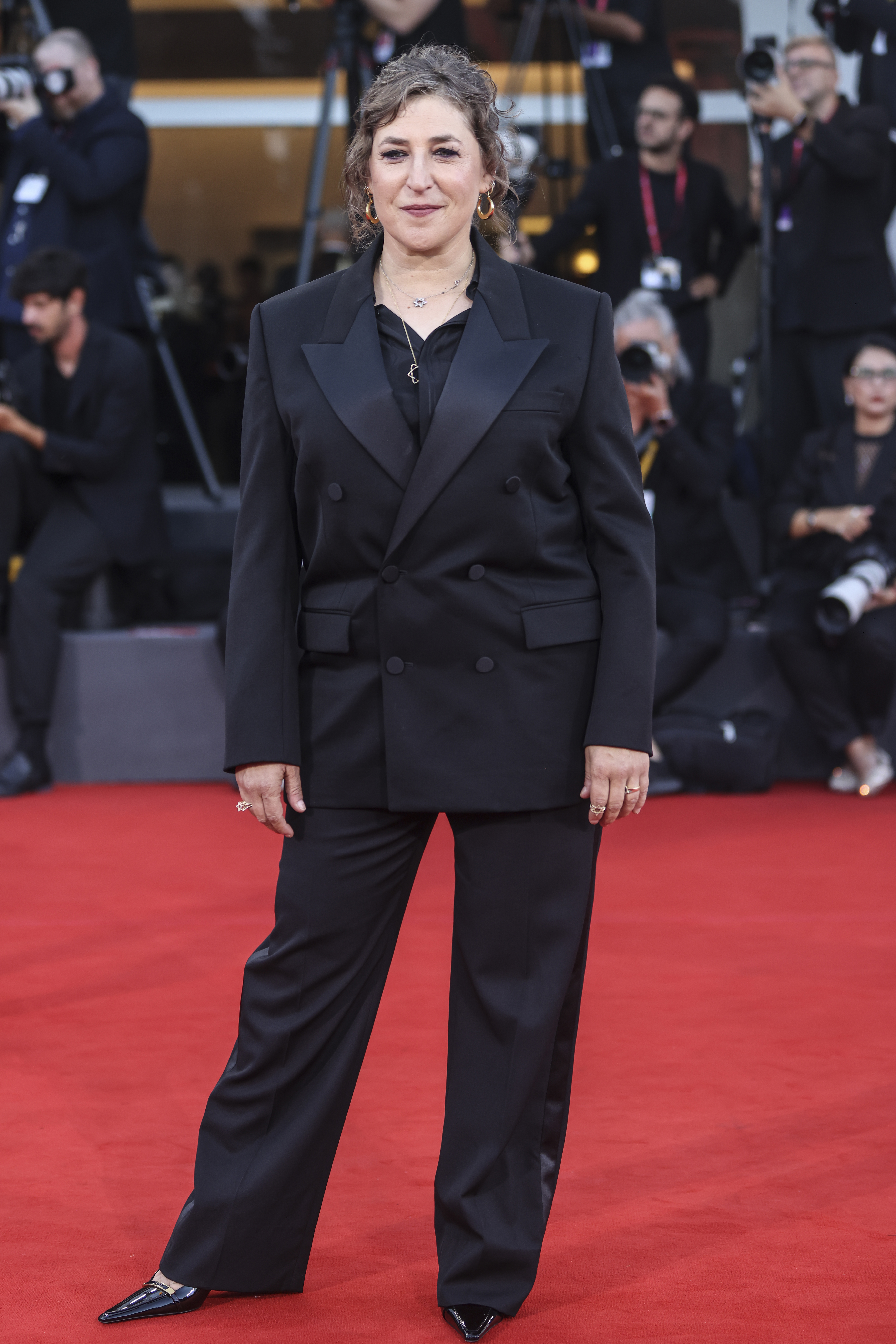Mayim Bialik on the red carpet at the 82nd Venice International Film Festival, wearing a black double-breasted suit.