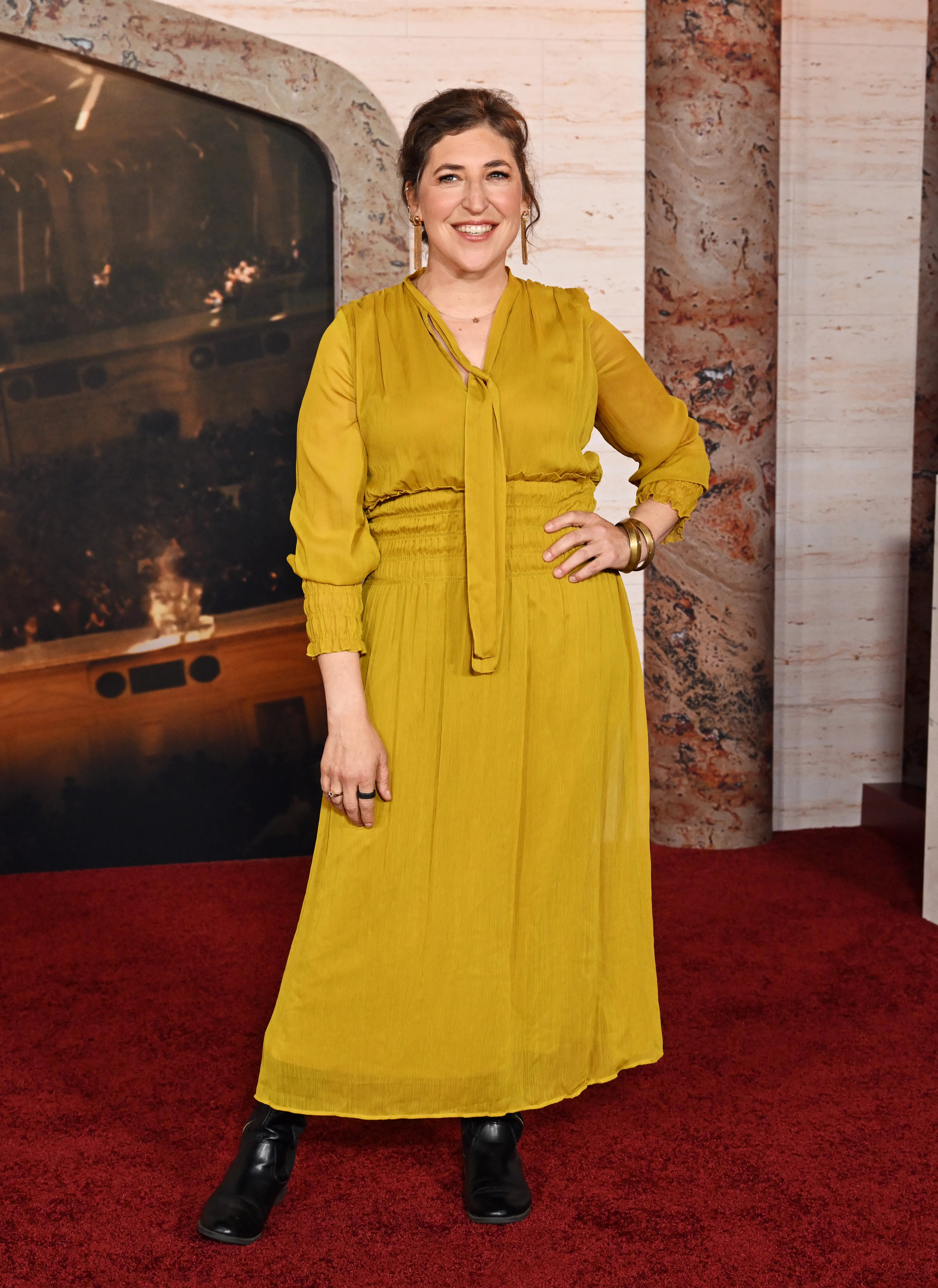 Mayim Bialik in a yellow dress and black boots on a red carpet.