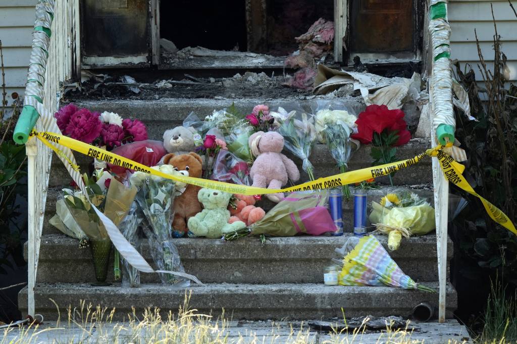 A memorial of flowers, candles, and teddy bears on the front steps of a burned-out home, with "FIRE LINE DO NOT CROSS" tape.