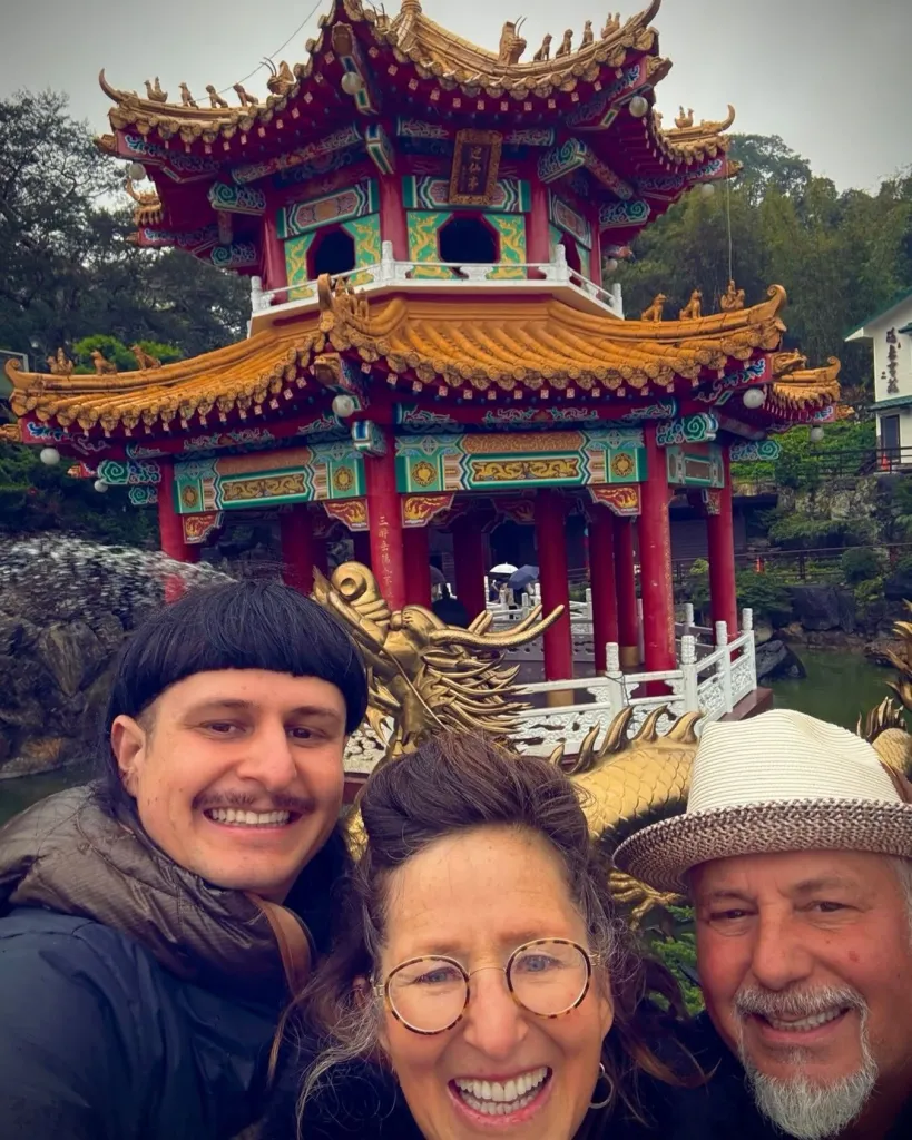 Three people posing for a selfie in front of a decorative red and gold pagoda-style building in Taipei.