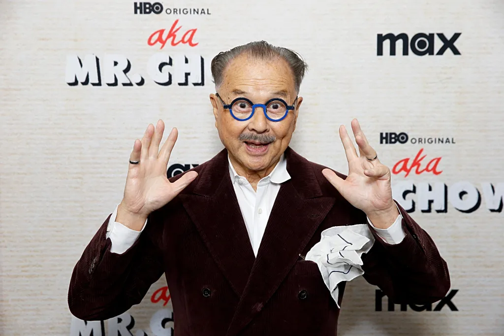 Michael Chow in a brown corduroy suit and blue-framed glasses, standing in front of an "AKA Mr. Chow" banner.