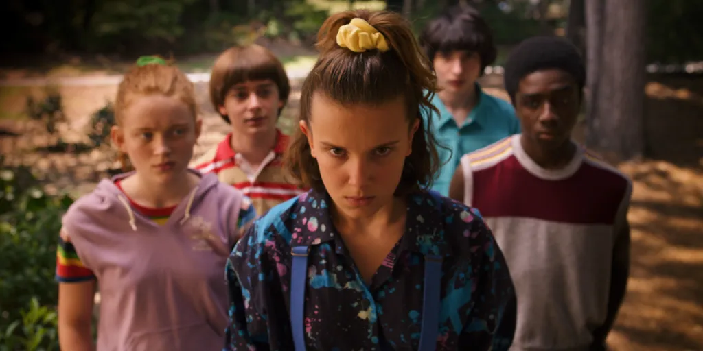 Millie Bobby Brown as Eleven with a serious expression, surrounded by Sadie Sink, Noah Schnapp, Finn Wolfhard, and Caleb McLaughlin, in a promotional photo for Stranger Things Season 3.