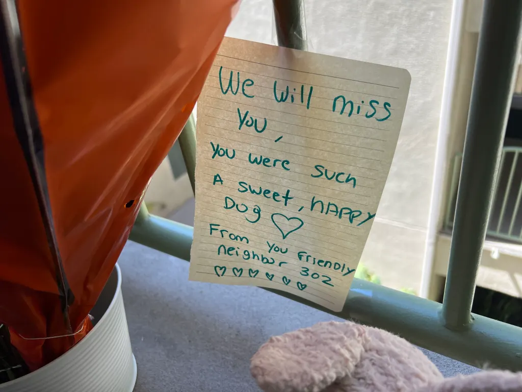 A handwritten note on lined paper attached to a railing reads, "We will miss you, You were such A sweet, HAPPY Dog ♡ From you friendly neighbor 302 ♡♡♡♡♡♡".