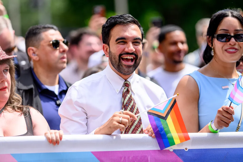 New York City Mayor Zohran Mamdani smiles at the 2026 NYC Pride March, holding a Progress Pride flag.