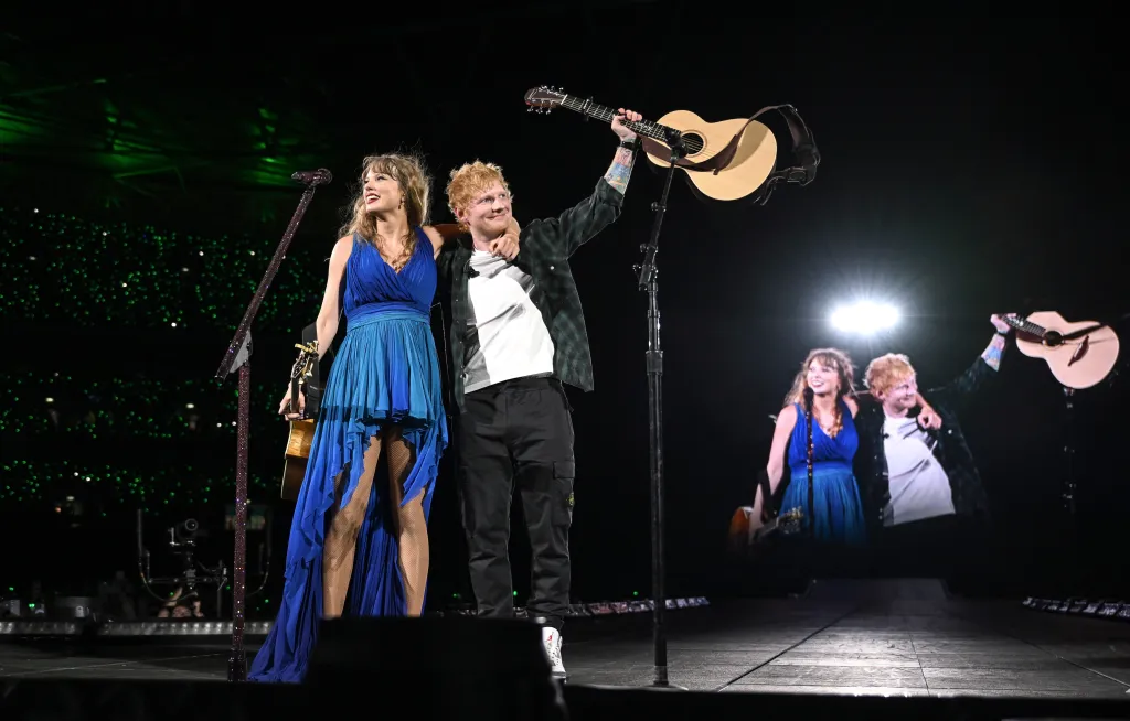 Taylor Swift in a blue dress and Ed Sheeran in a plaid shirt and black pants perform on stage with guitars, with a large screen behind them showing the same image.