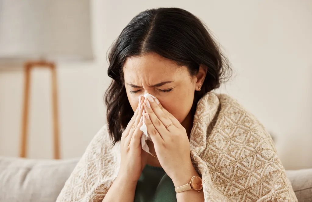 A woman with dark hair, wrapped in a blanket, blows her nose into a tissue.