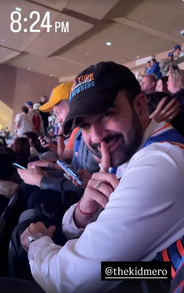 Zohran Mamdani wearing a black "New York Knicks Nowhere" baseball cap, finger to his lips, at a New York Knicks game.