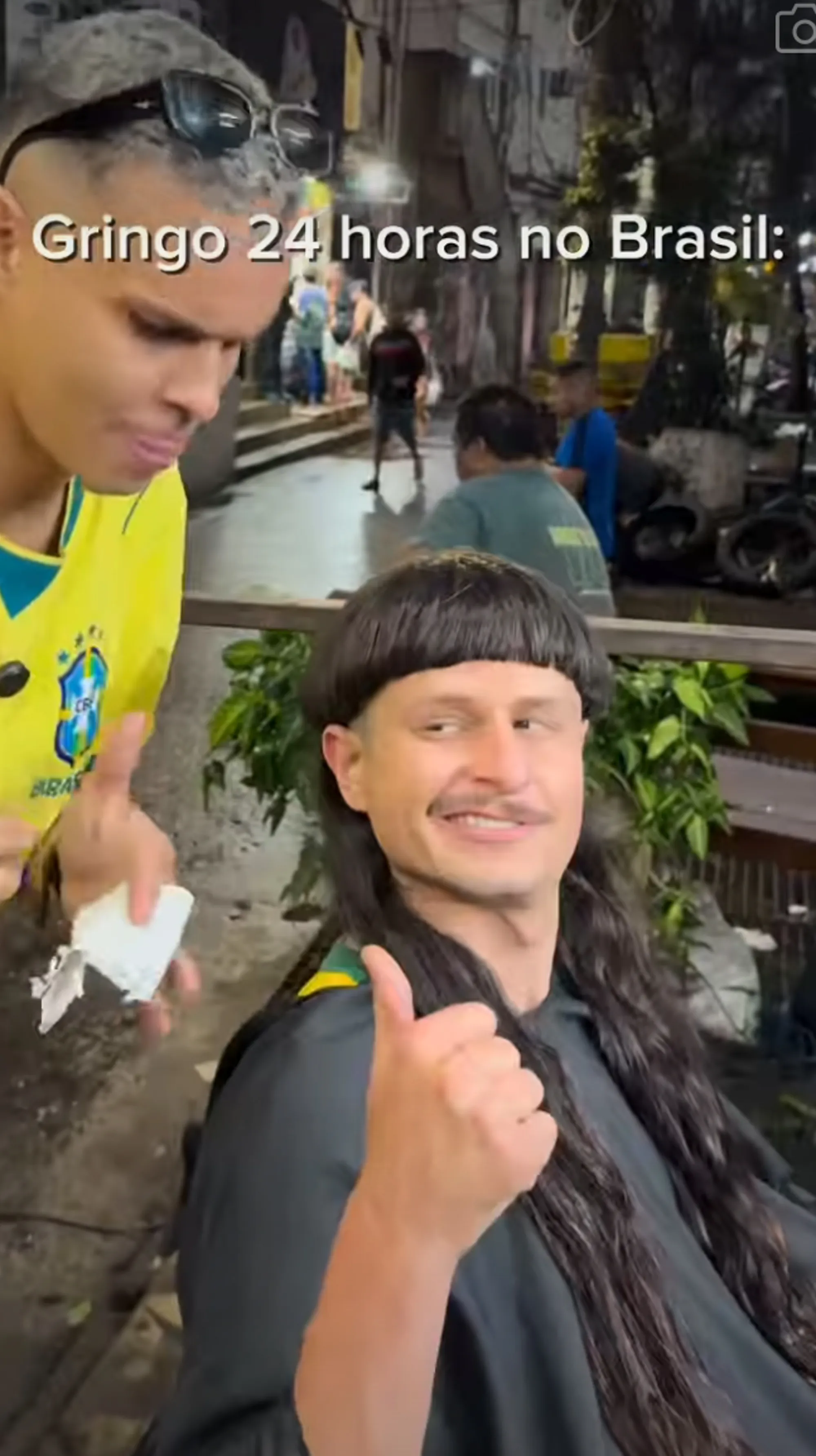 A man with a mullet haircut and a mustache gives a thumbs up while another man in a Brazil soccer jersey shows him something.