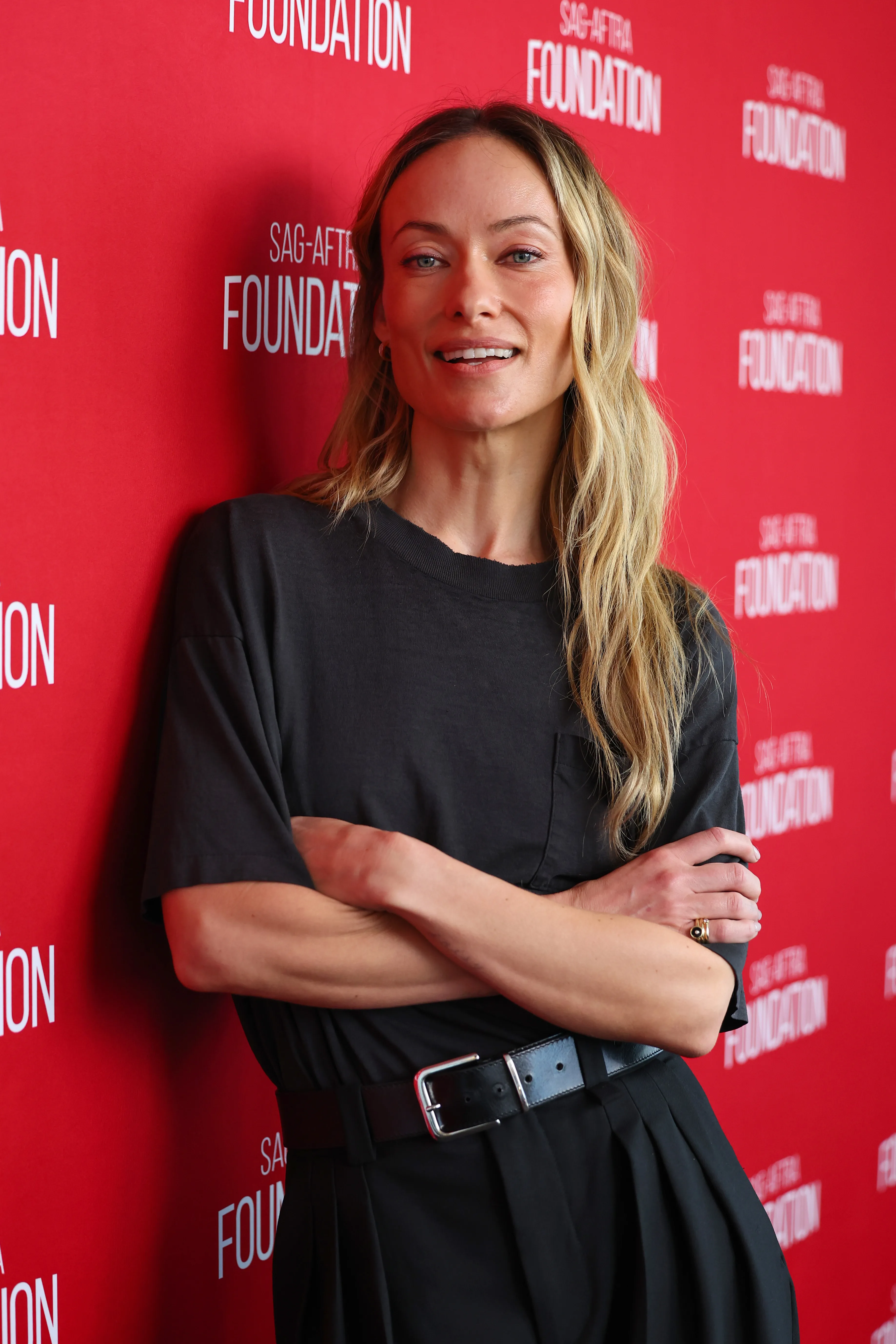 Olivia Wilde smiling with her arms crossed in front of a red backdrop with "SAG-AFTRA Foundation" printed on it.