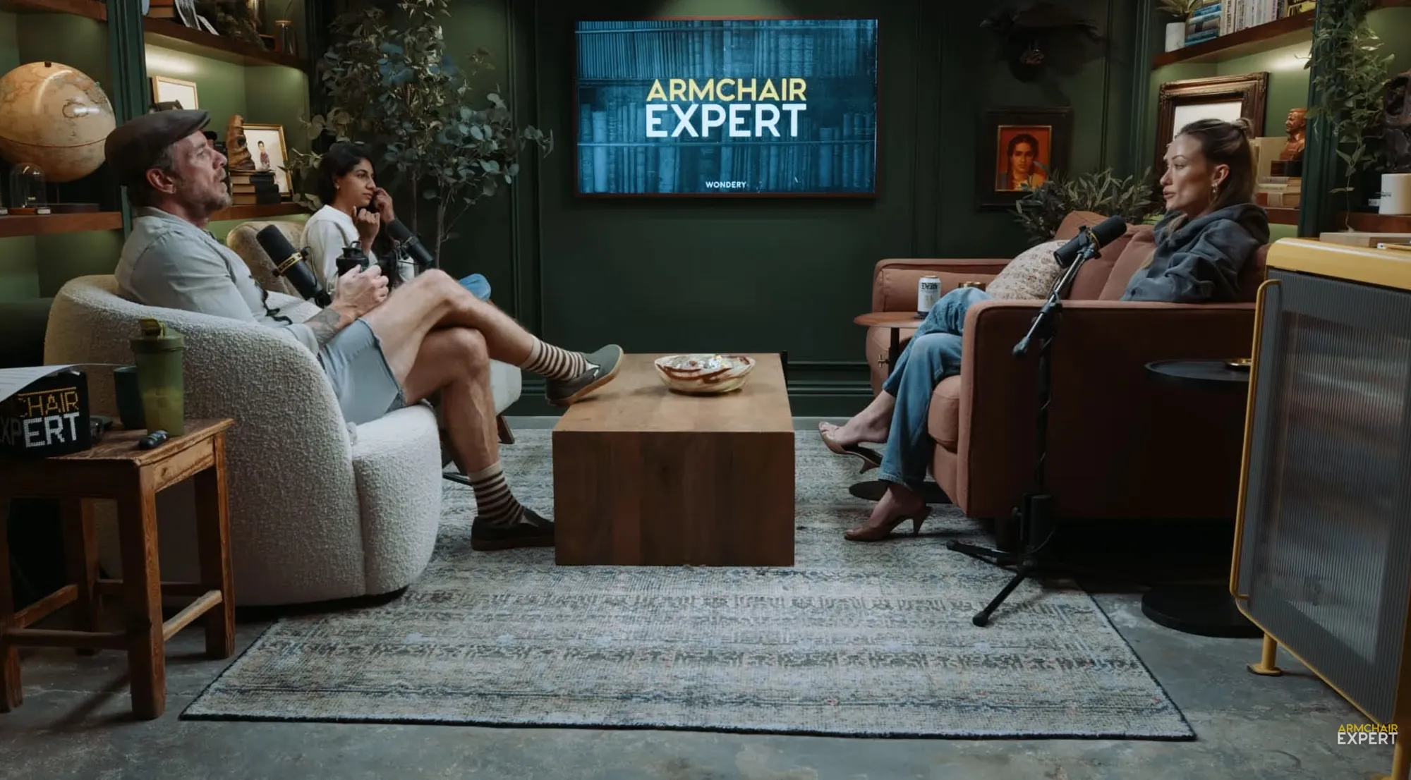 Dax Shepard, a woman with dark hair, and Olivia Wilde seated in a living room set for an Armchair Expert podcast.