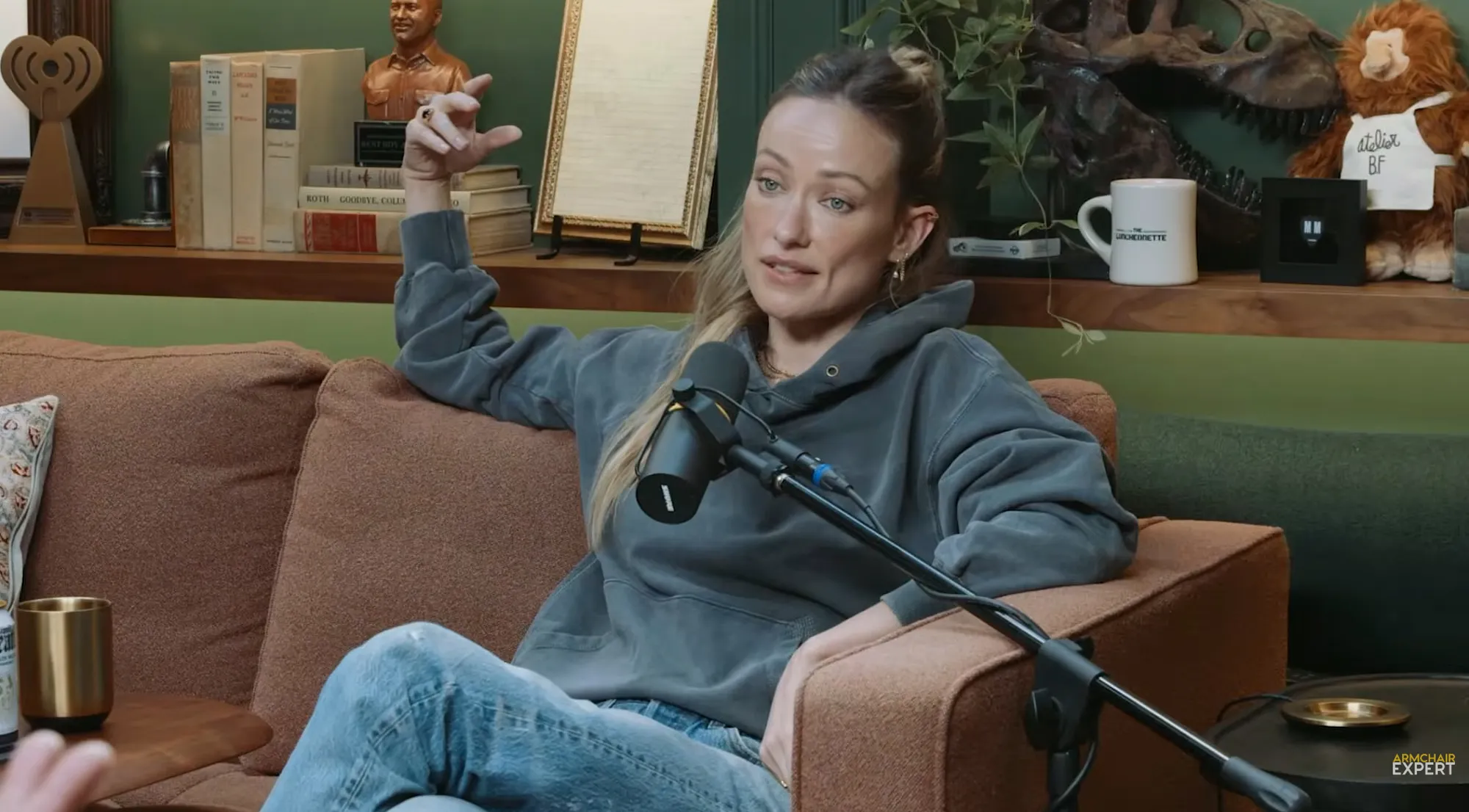 Olivia Wilde on a podcast, sitting on a brown couch with a microphone in front of her.