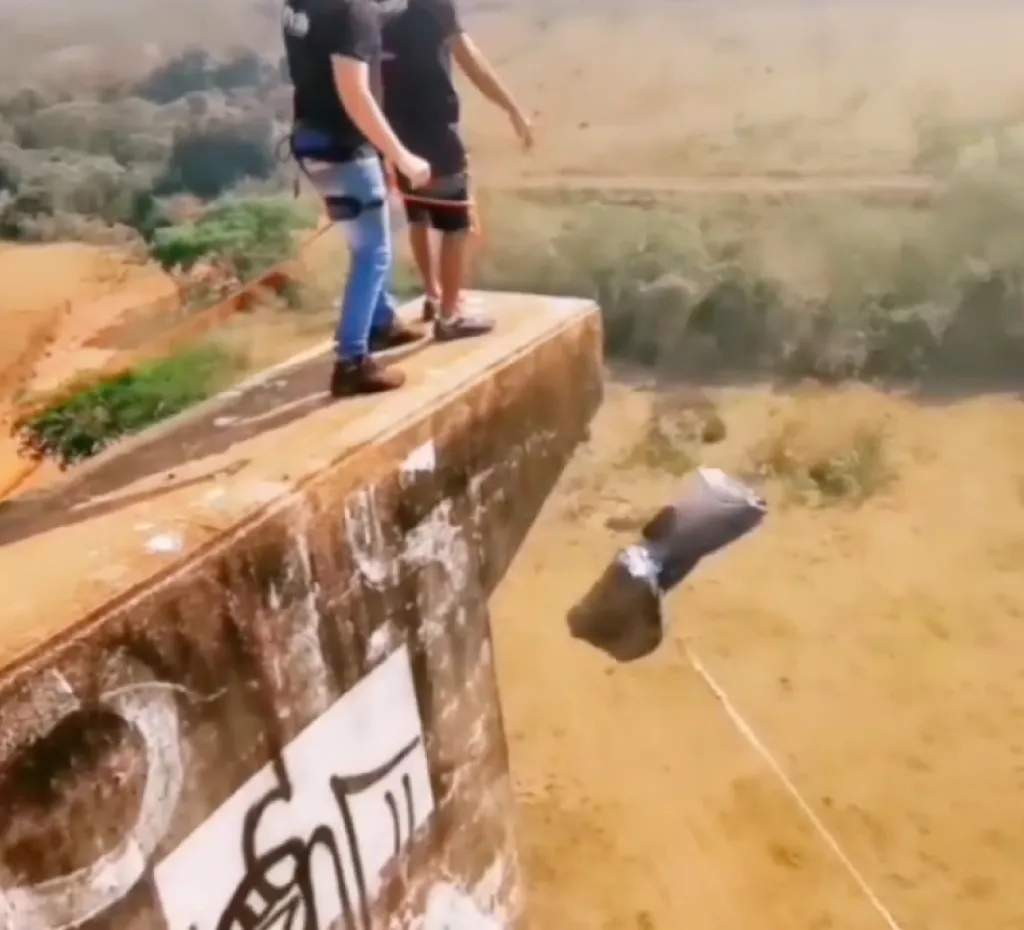 Two men on a concrete ledge preparing for a rope jump, with one man already secured with a harness and rope.