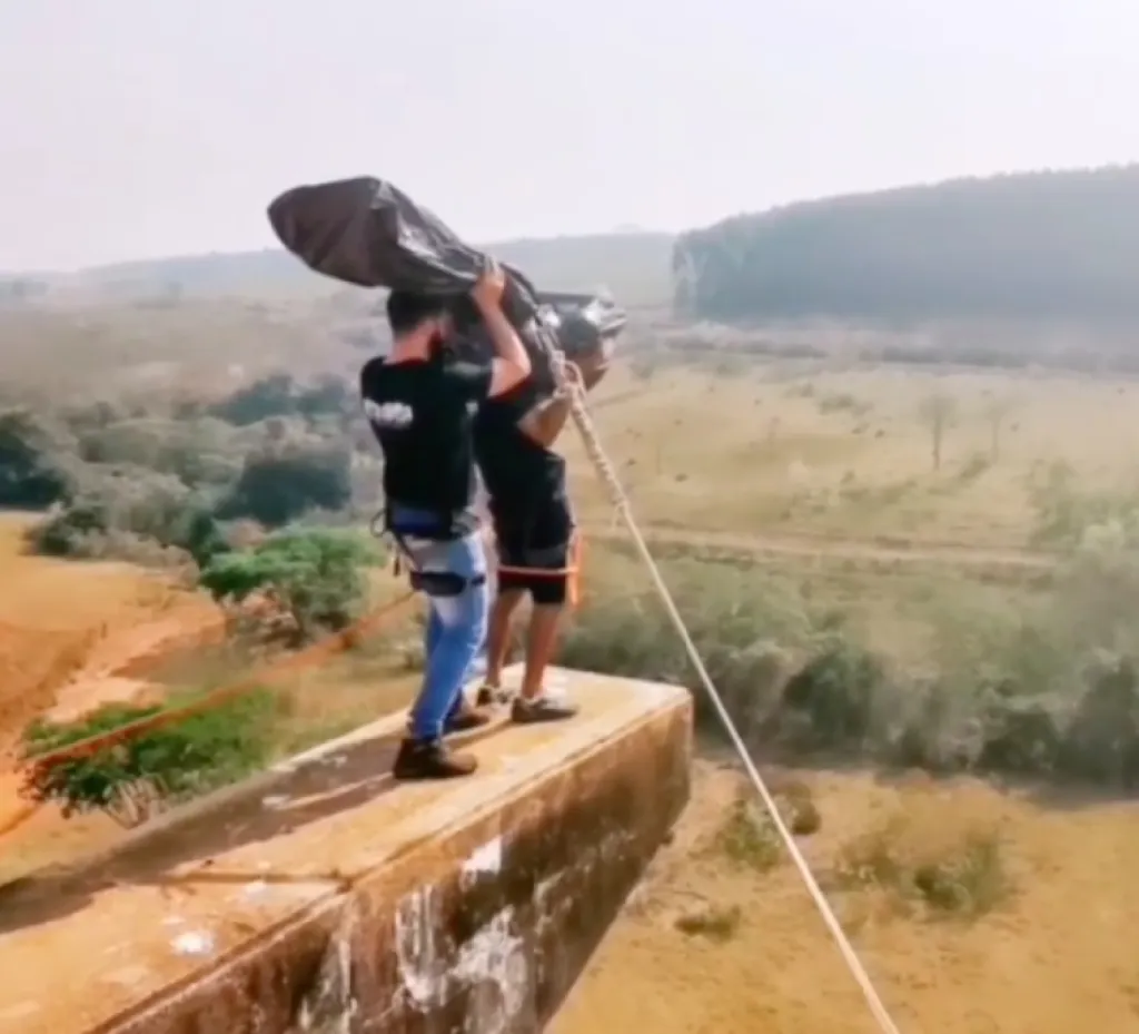 Two men standing on a concrete ledge, preparing for a rope jump, with a wide, hilly landscape in the background.
