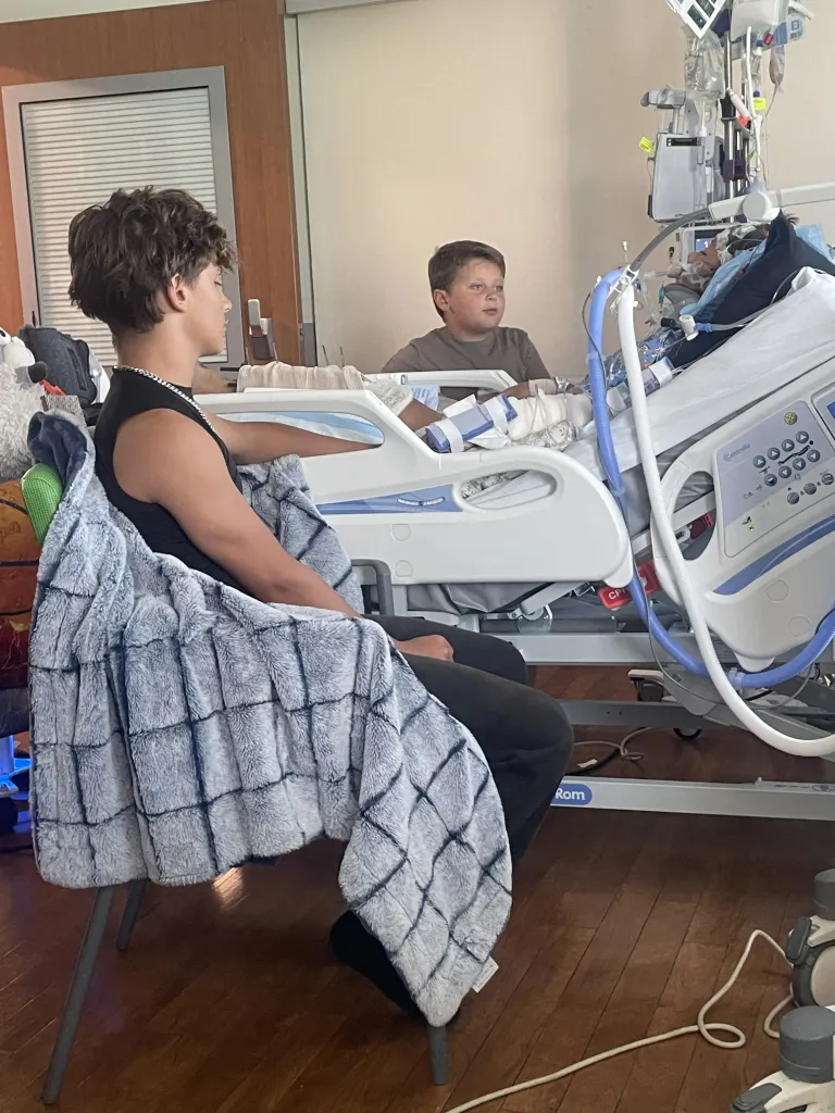 Two boys in a hospital room, one sitting in a chair and the other in a hospital bed with medical equipment.