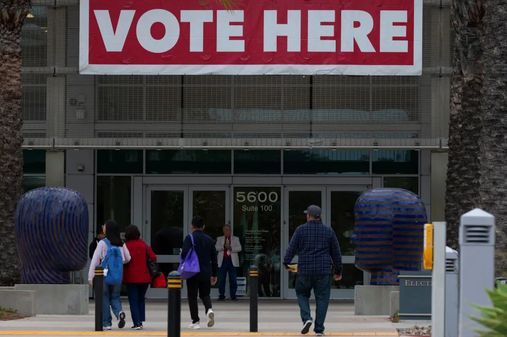 People arrive at a California primary election polling place with a "VOTE HERE" banner.