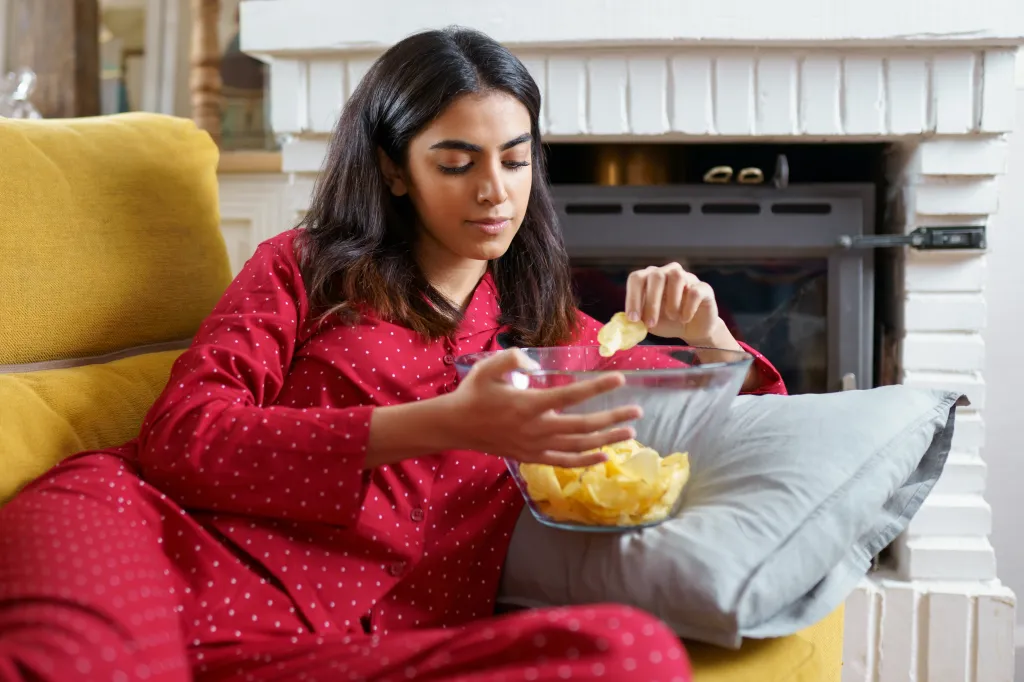 A woman in red pajamas watches TV while eating potato chips.