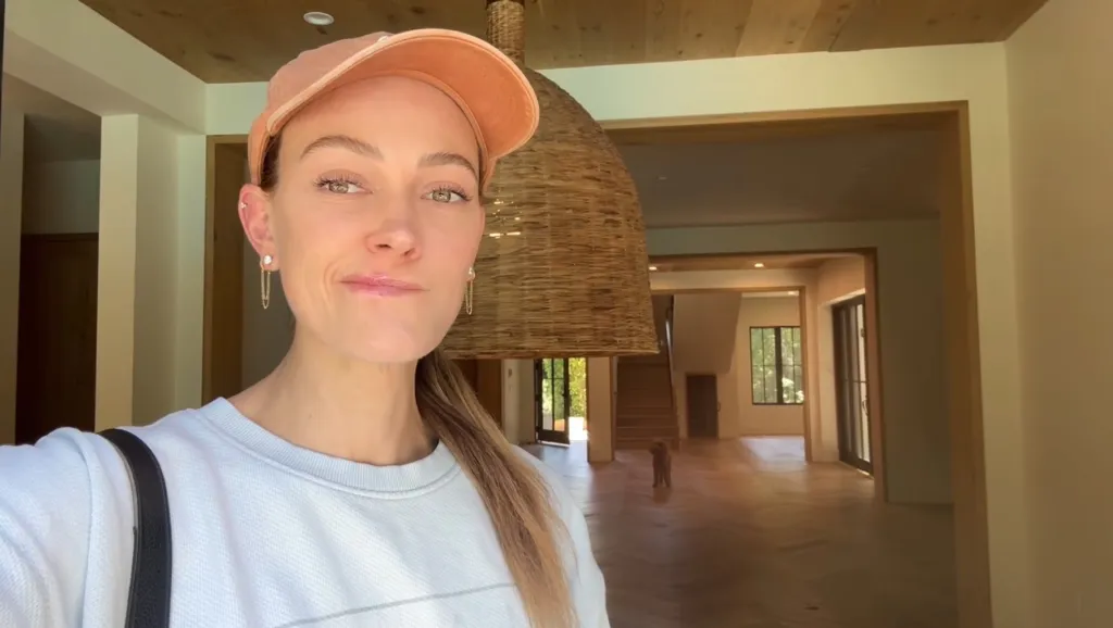 Peta Murgatroyd in a peach cap and white sweatshirt, standing inside a large, empty house with hardwood floors and a woven pendant light.