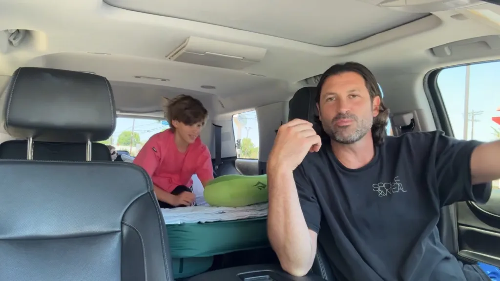 Maksim Chmerkovskiy and Shai sitting in a car with an inflatable mattress in the back.