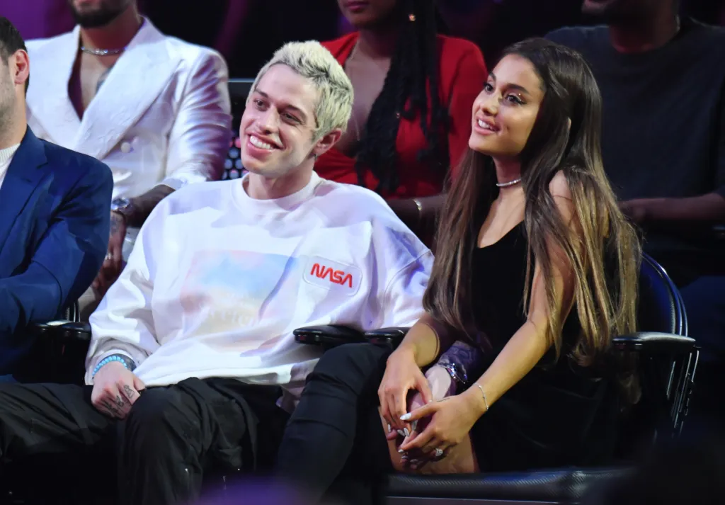 Pete Davidson, in a white sweatshirt, and Ariana Grande, in a black dress, sitting together at an awards ceremony.