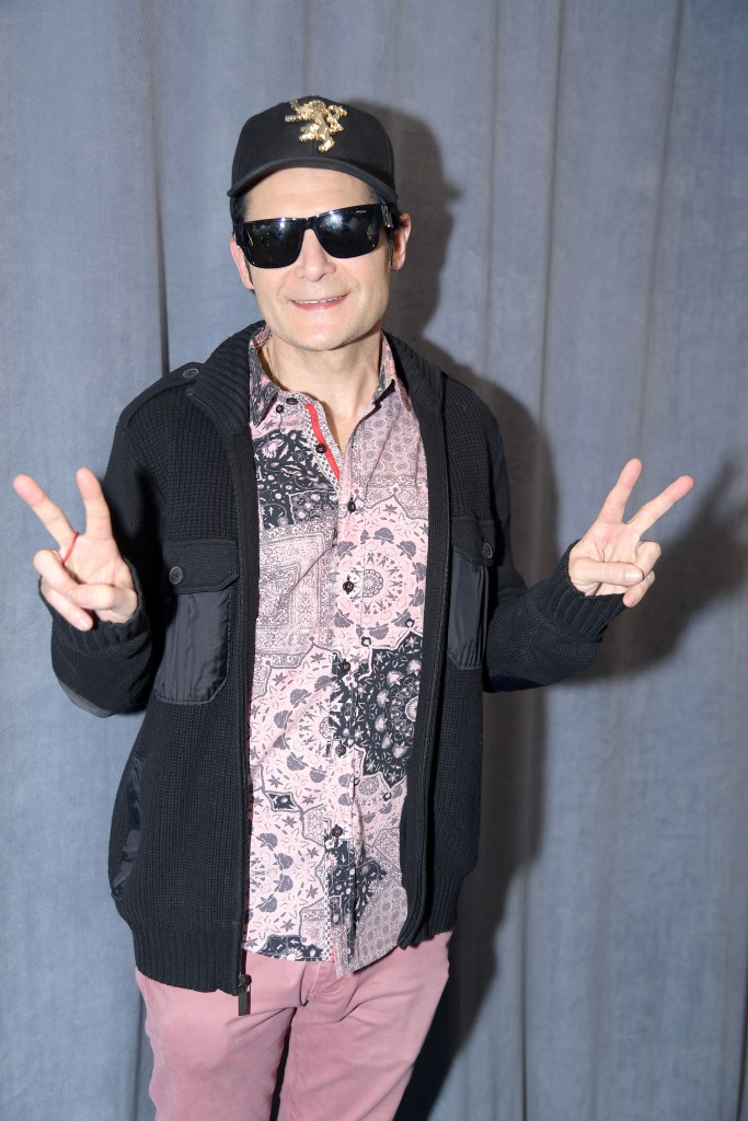 Corey Feldman wearing a black hat, sunglasses, a patterned shirt, a black cardigan, and pink pants, making peace signs with both hands.