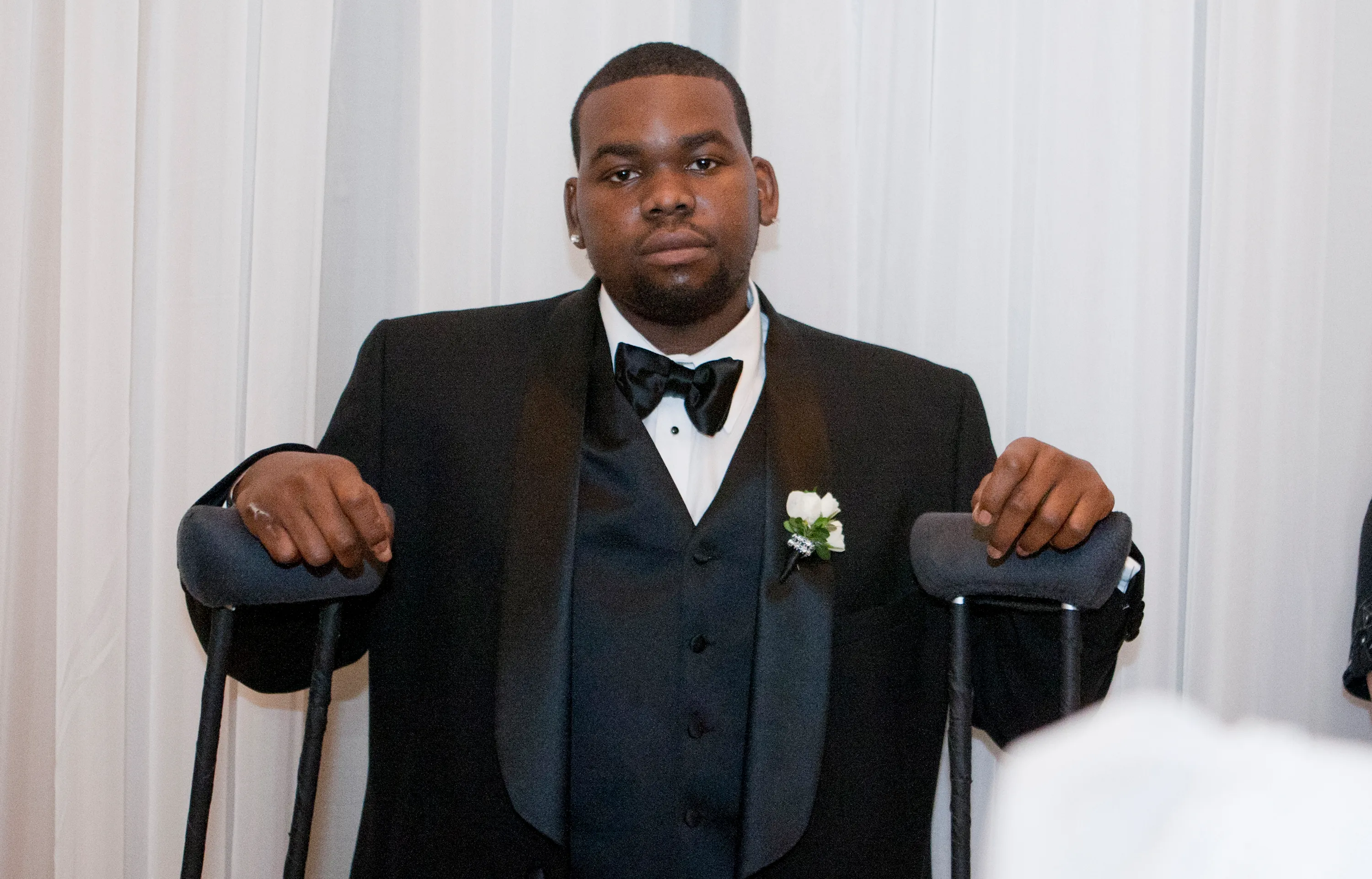 Bryson Bryant from "I Dream of NeNe: The Wedding" stands in a tuxedo, holding onto crutches, looking at the camera.