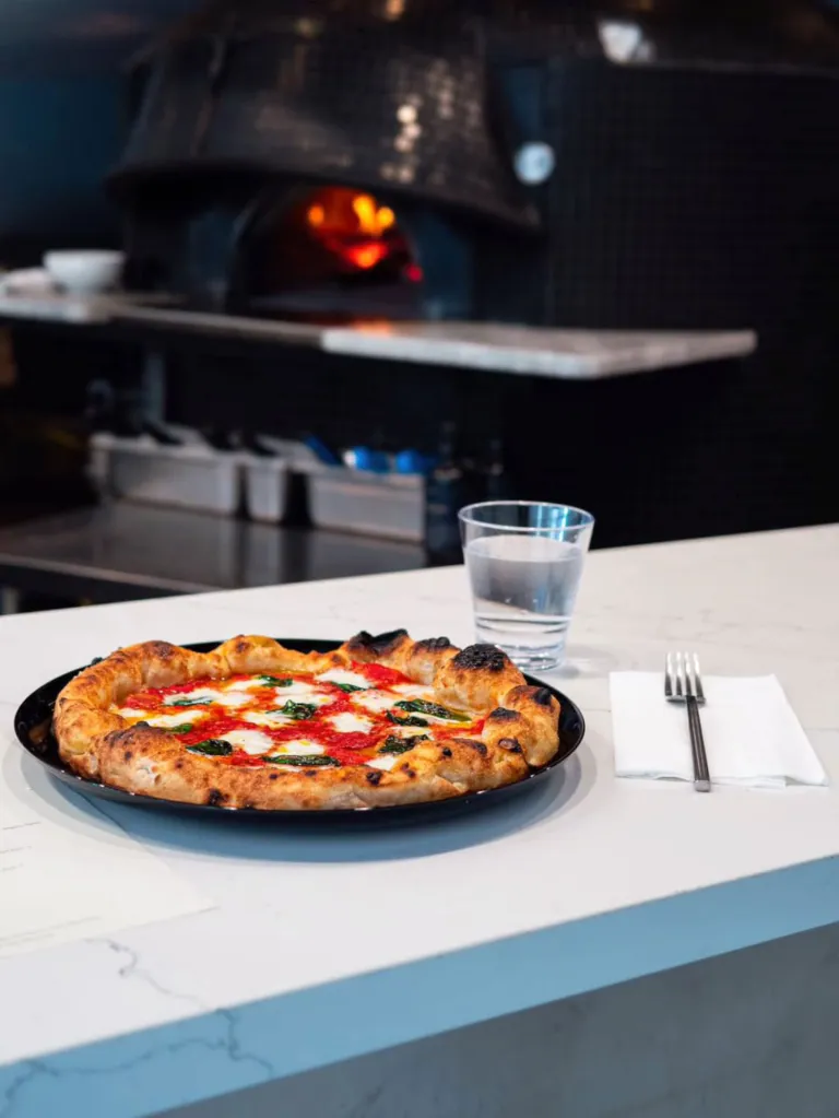 A freshly baked Pizza Sei pizza with mozzarella, tomato sauce, and basil on a white counter with a glass of water and a fork, with a pizza oven in the background.