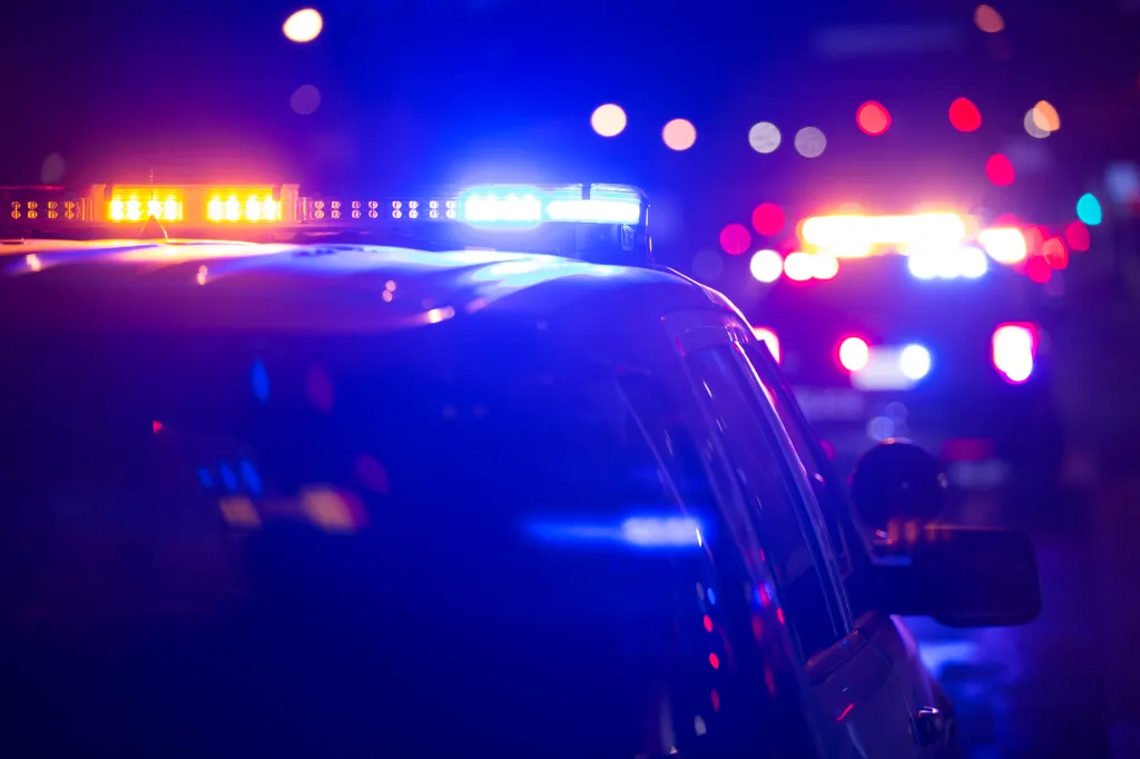 Police car with flashing blue and orange lights at night, with other emergency vehicle lights in the background.
