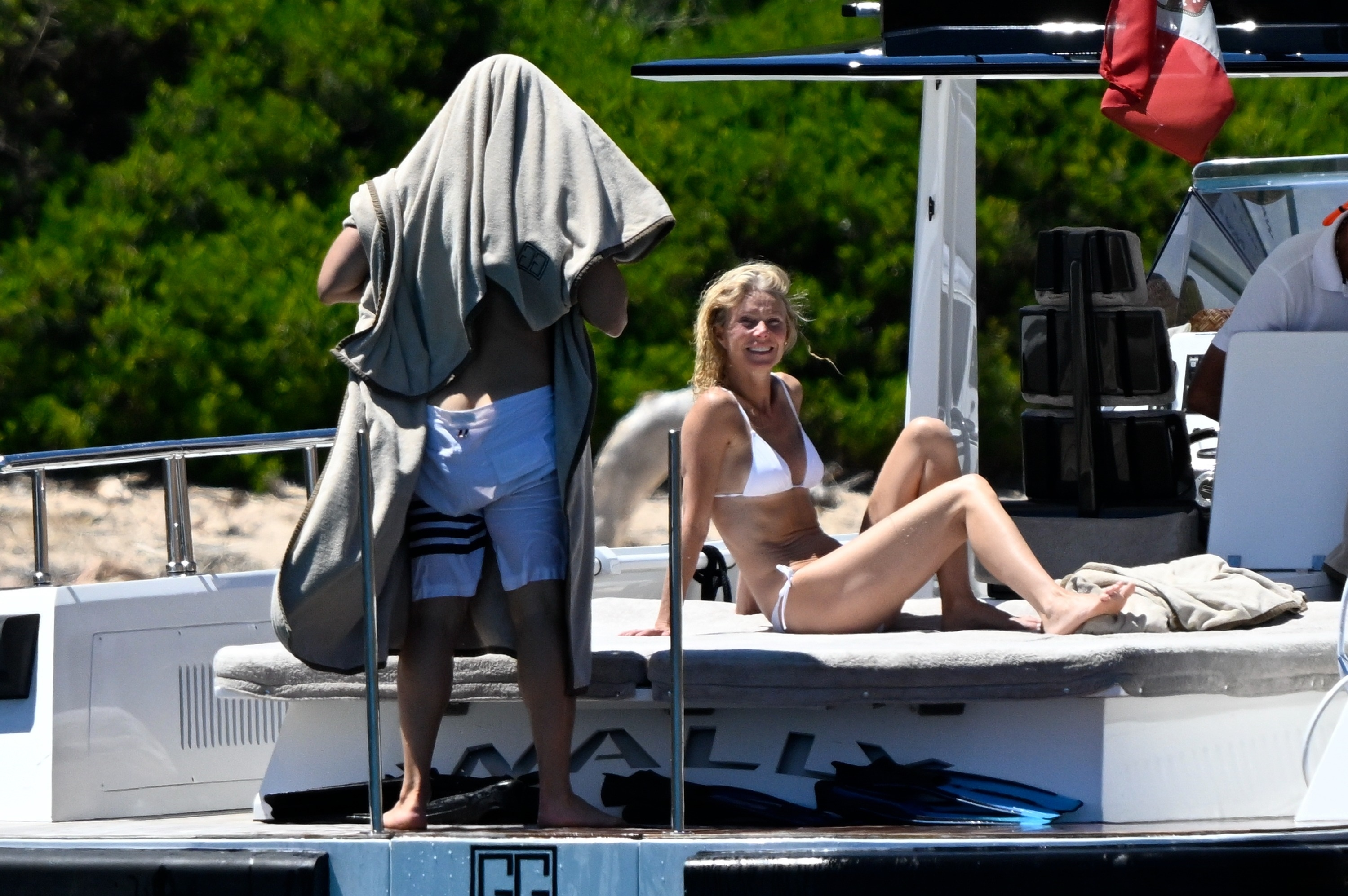 Gwyneth Paltrow in a white bikini sits on a yacht while Brad Falchuk, wrapped in a towel, stands beside her.