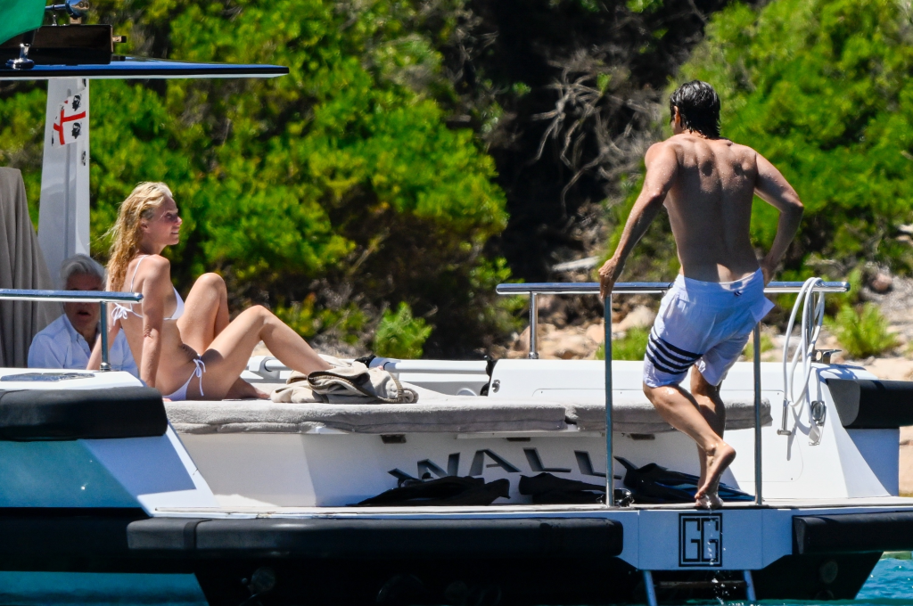 Gwyneth Paltrow in a white bikini sits on a yacht as Brad Falchuk in white swim trunks steps onto the boat.