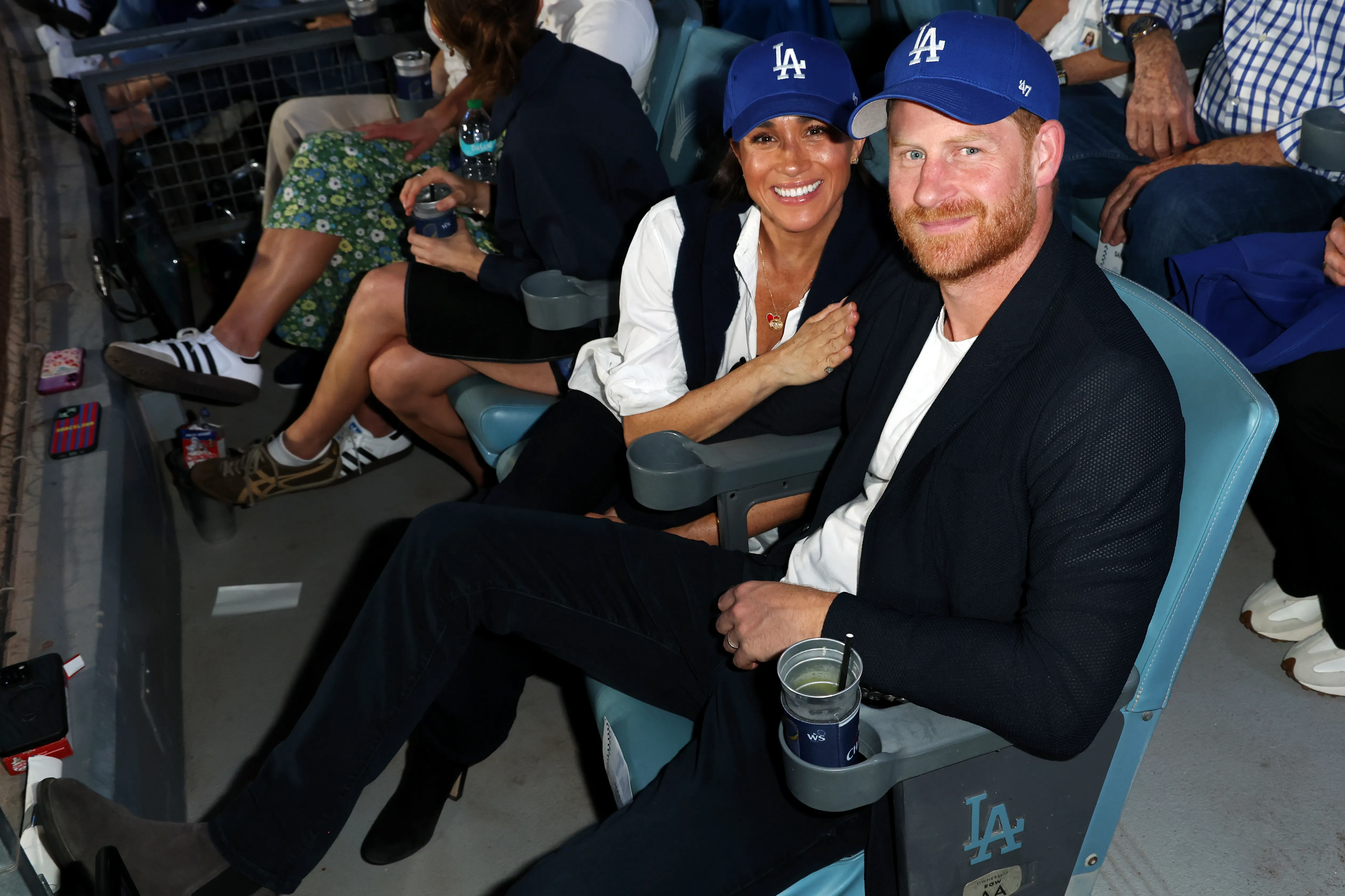 Meghan, Duchess of Sussex and Prince Harry, Duke of Sussex, wearing blue "LA" baseball caps, smile while seated at Dodger Stadium.