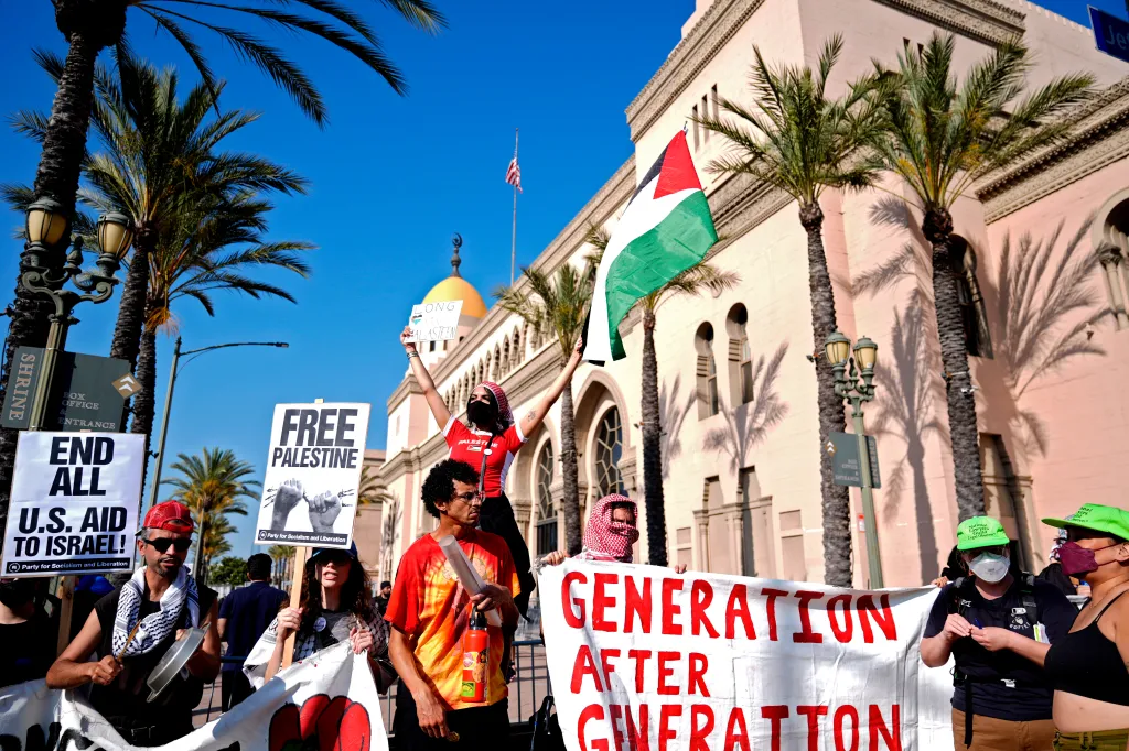 Pro-Palestinian demonstrators rally outside the Shrine Auditorium, holding signs saying "END ALL U.S. AID TO ISRAEL!" and "FREE PALESTINE", with one person holding a Palestinian flag.