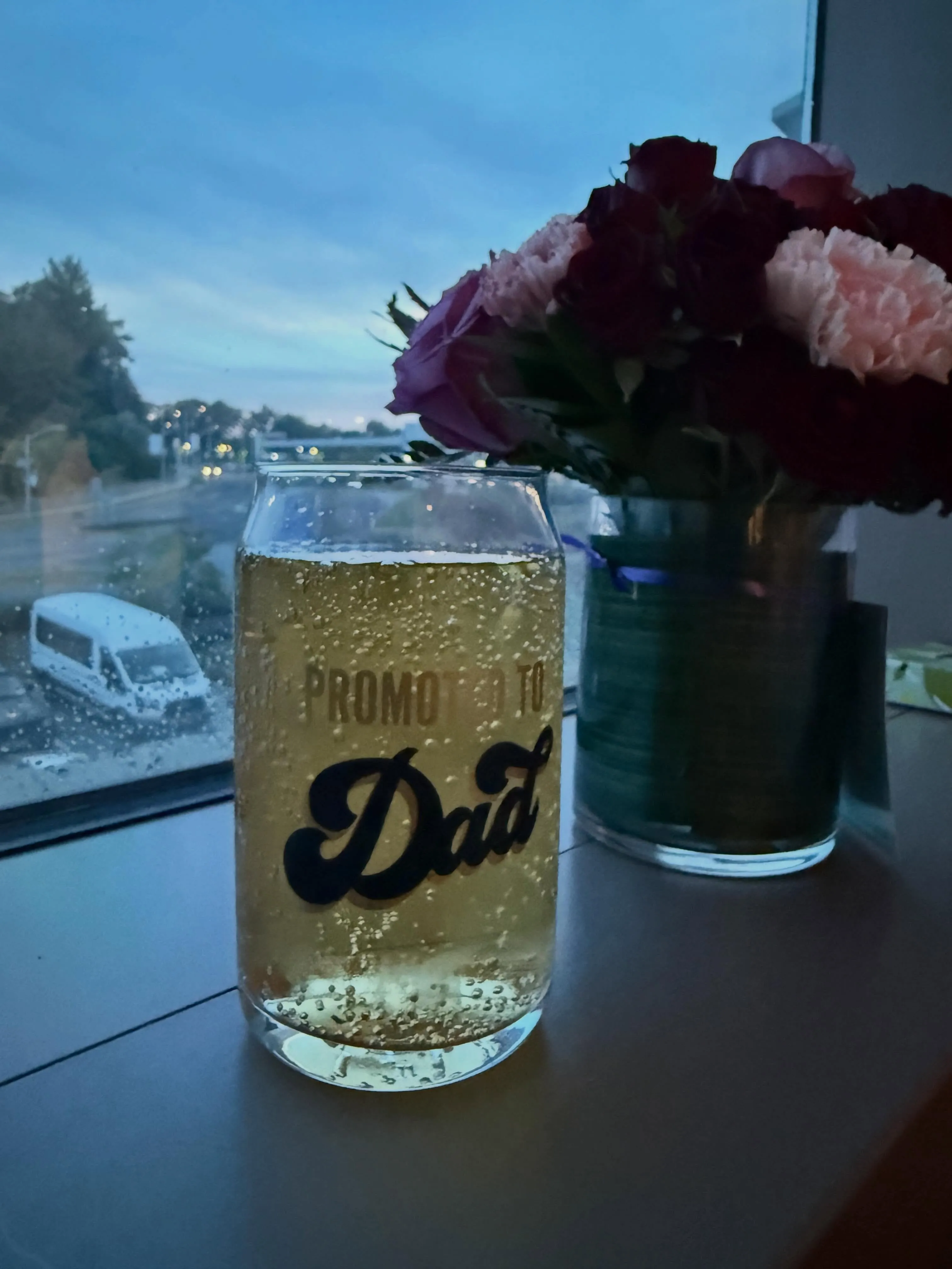 A glass with a "Promoted to Dad" decal, filled with a sparkling beverage, next to a vase of flowers, with an out-of-focus city view outside a window.