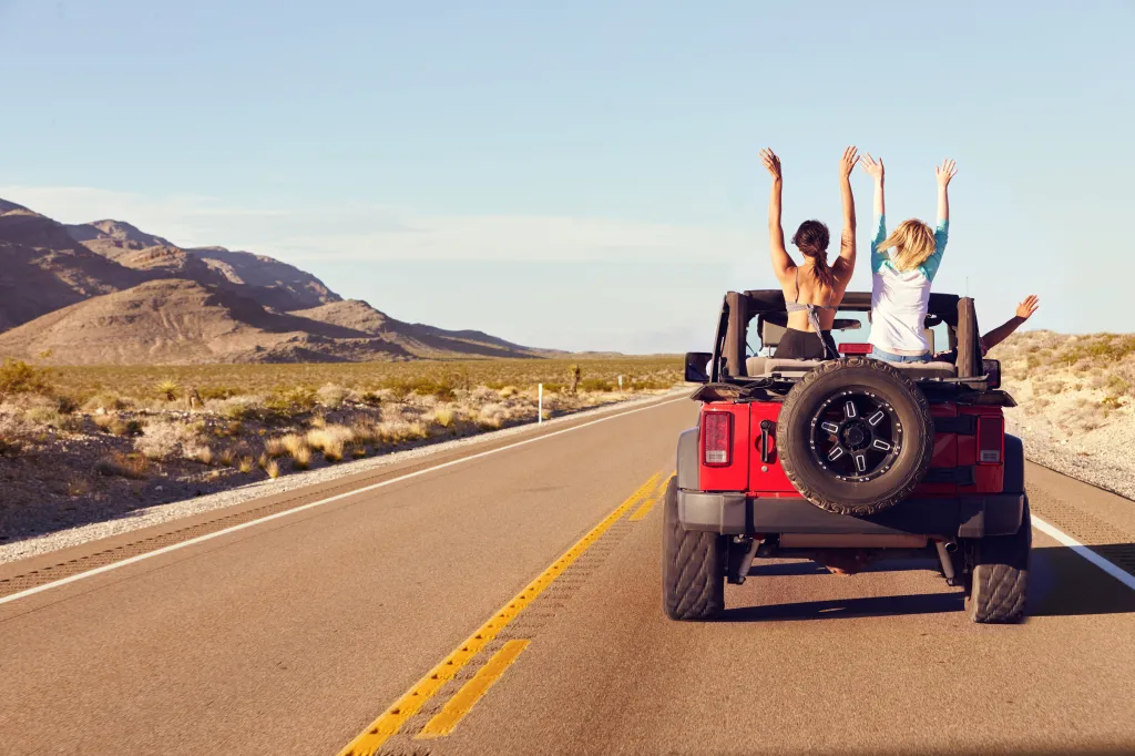 Rear view of four friends on a road trip, driving in a convertible car with their arms raised.