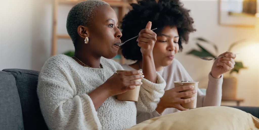 Two Black women sitting on a couch, eating ice cream from paper cups.