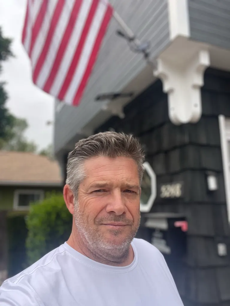 Richie Varga in a white shirt, with an American flag and a house in the background.