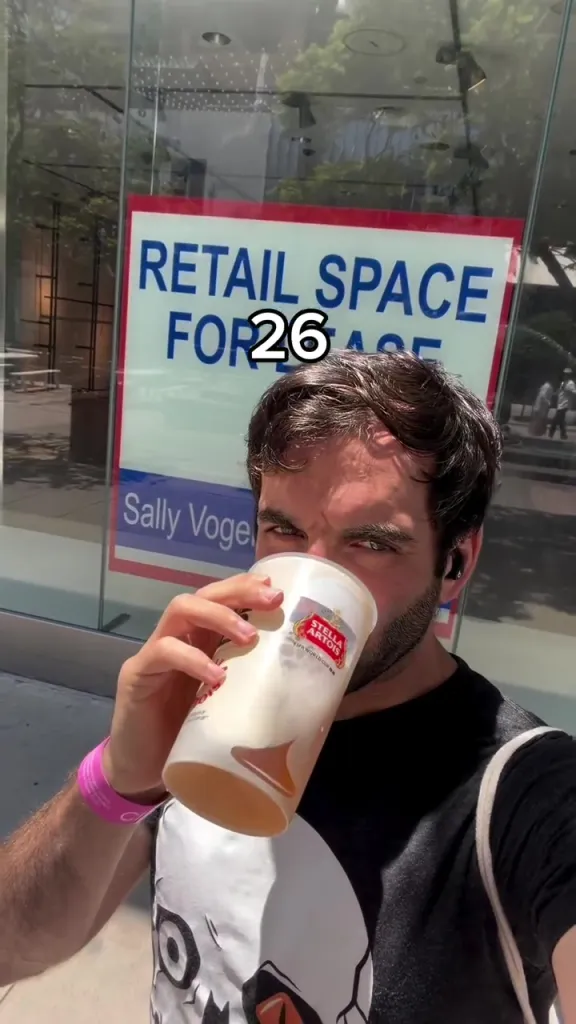 A man drinking from a large cup in front of a "Retail Space for Lease" sign with the number 26 overlaid on it.