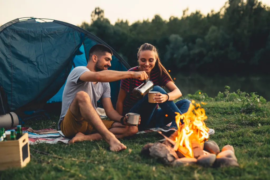 Couple by a campfire pouring coffee into mugs.