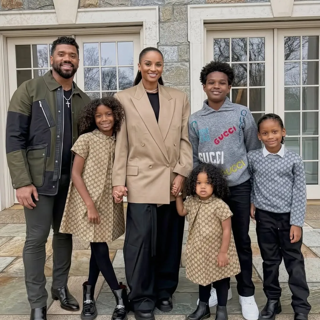 Russell Wilson and Ciara with their children, including a young girl and toddler, holding hands in front of a stone house.