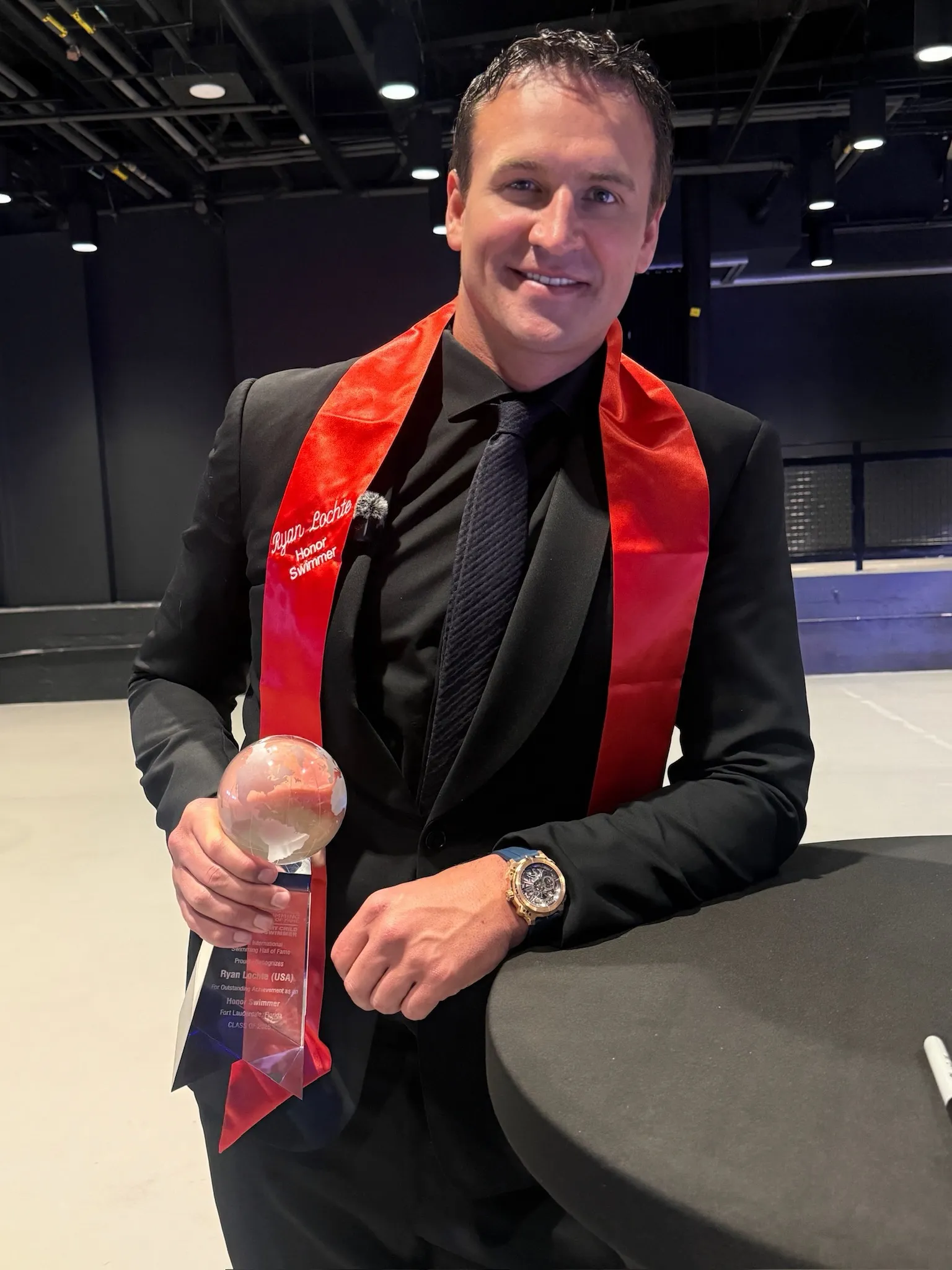 Ryan Lochte wearing a red sash that says "Ryan Lochte Honor Swimmer" and holding a trophy from the International Swimming Hall of Fame.