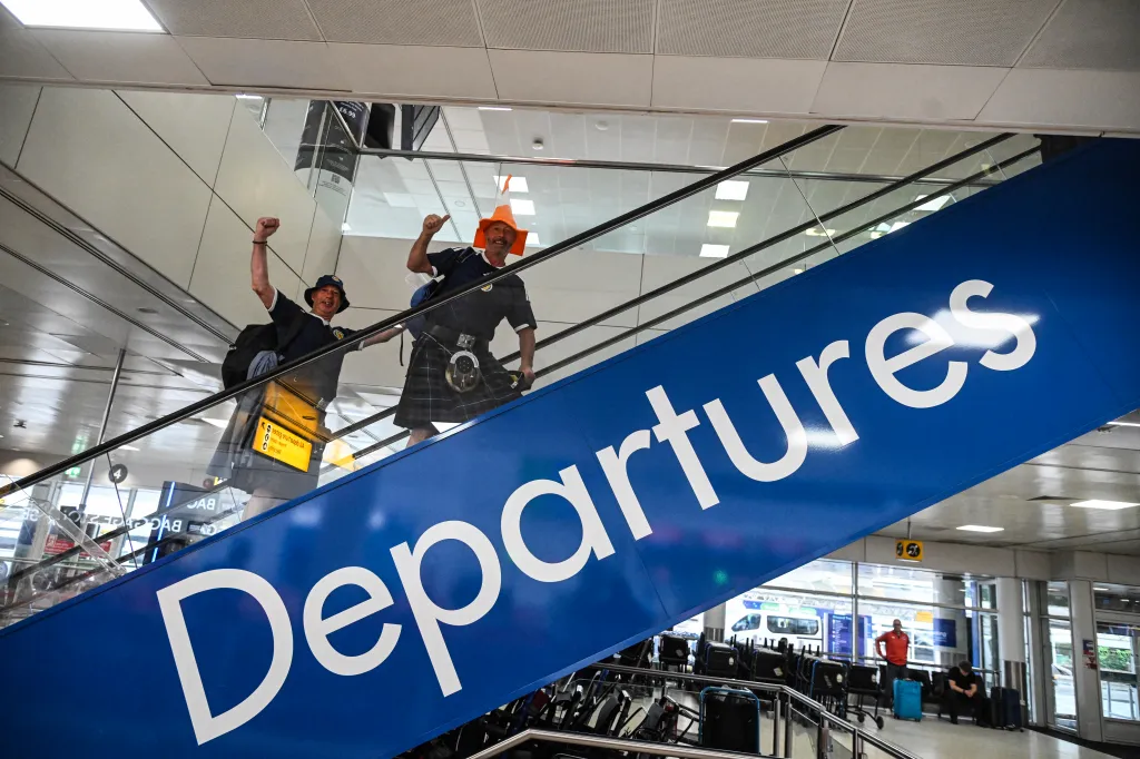 Two jubilant Scotland football fans on an escalator under a "Departures" sign at Glasgow Airport.
