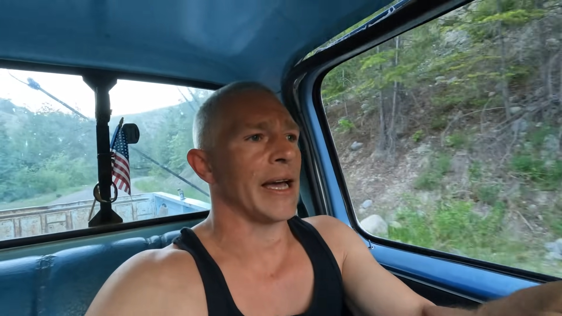 A man in a blue truck with an American flag attached to the back window.