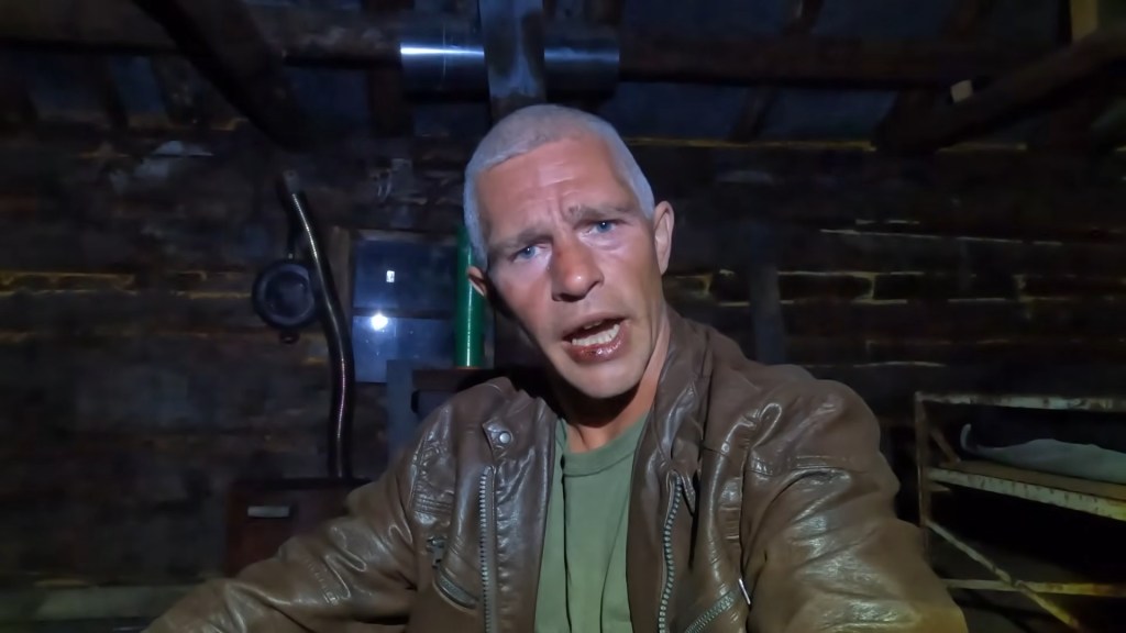 Matt Brown, an alum of "Alaskan Bush People," wearing a brown leather jacket, with dark gray hair, appears to be speaking to the camera in a dimly lit, rustic room with wooden walls.