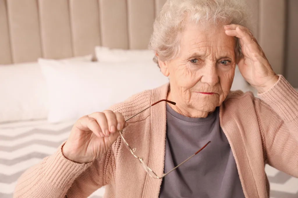 Senior woman with a headache in her bedroom, holding her glasses.