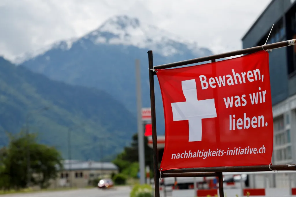 A red sign with a white Swiss cross and the German text "Bewahren, was wir lieben" (Preserve what we love) and "nachhaltigkeits-initiative.ch" in front of a snowy mountain in Glarus, Switzerland.