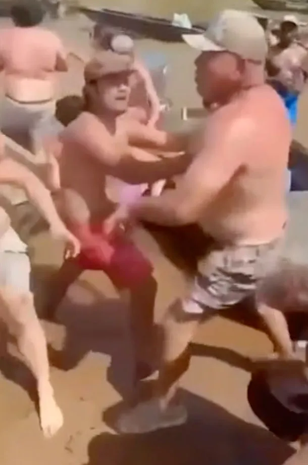 A large fight with multiple shirtless men on a sandy riverbank.