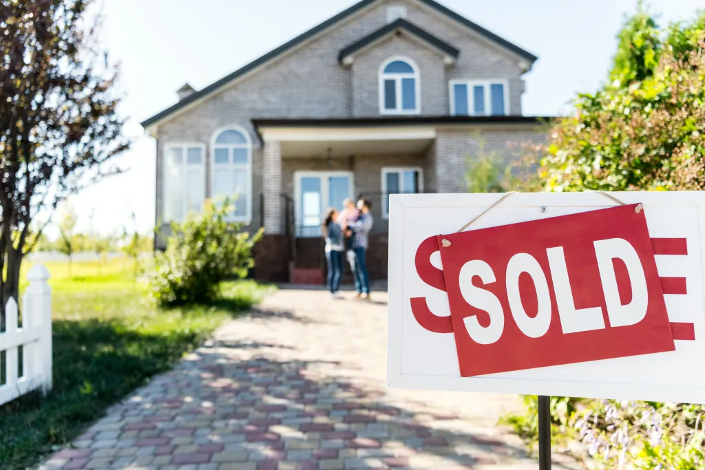 A "SOLD" sign in front of a new house, with a family visible in the background.