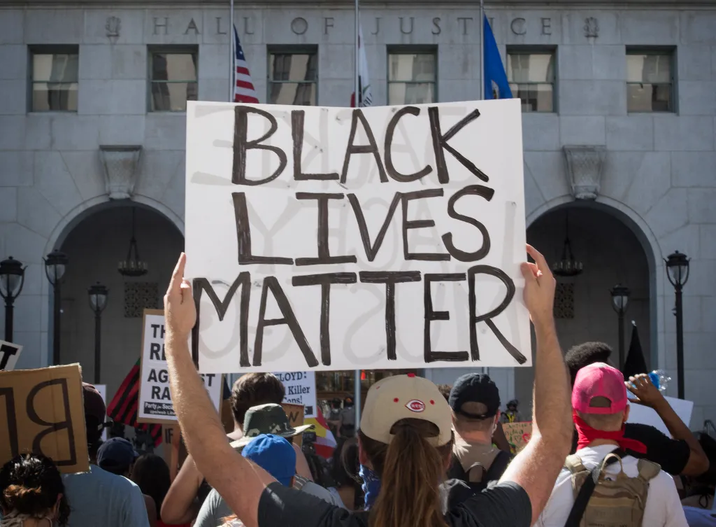 A person at a Black Lives Matter protest holds up a sign that reads "BLACK LIVES MATTER" in front of the Hall of Justice building.