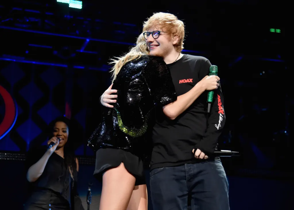 Taylor Swift embracing Ed Sheeran on stage, with Swift in a black sequined jacket and Sheeran in a black long-sleeved shirt holding a green microphone.